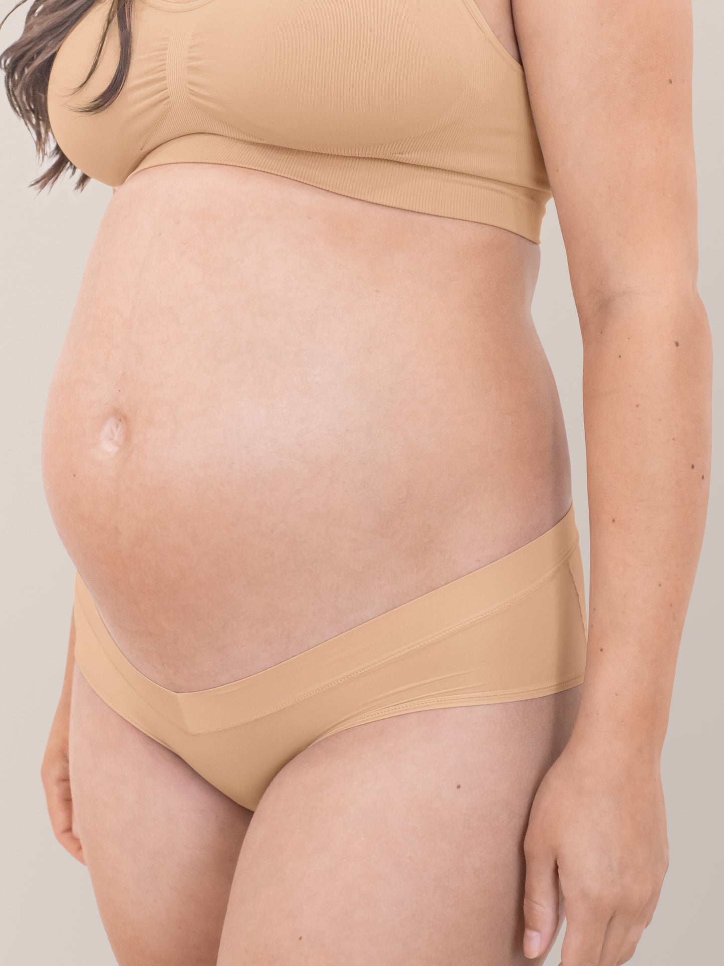 Closeup of a pregnant model wearing the Grow with Me™ Maternity & Postpartum Hipster in Beige with her hands at her sides.