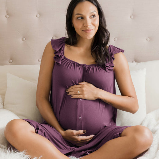 Model sitting on a bed while wearing the Ruffle Strap Labor & Delivery Gown in Burgundy Plum with her hands on her pregnant belly. @model_info:Julana is 5'9